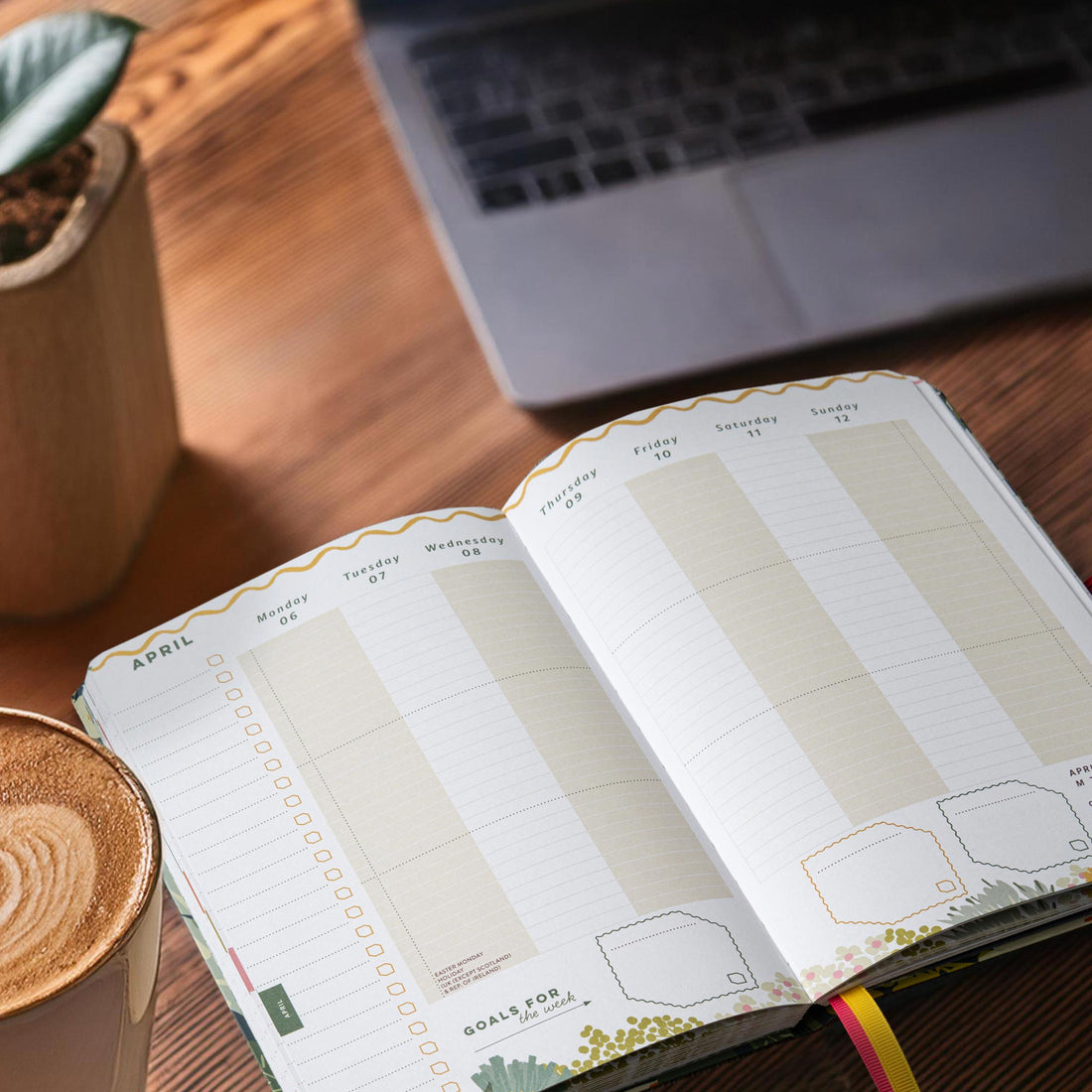 Open academic diary with vertical layout on a desk next to a coffee and a plant in a stone pot. 