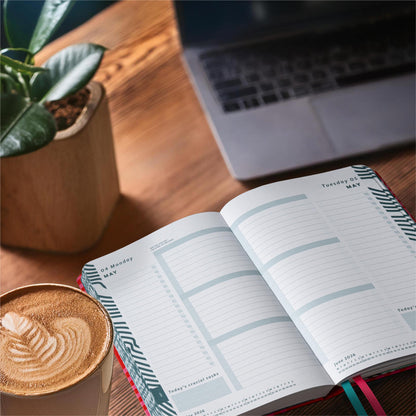 Open mid year diary, with page a day layout on a desk next to a coffee and a plant in a stone pot. 